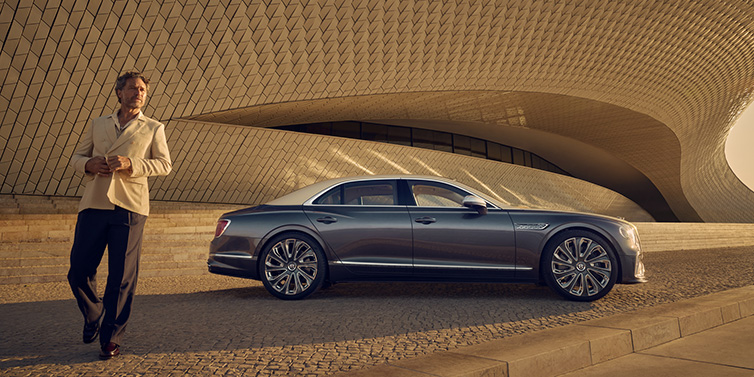 Bentley Sydney Bentley Flying Spur Mulliner sedan in Rose Gold over Magnetic duotone paint parked by a curved wall with person in suit jacket standing on the left